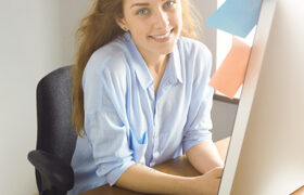 Portrait of cute female designer working at home on new ideas. Redhead young woman sitting at the wooden table in casual blue shirt looking with happy smile at the camera, with laptop in front of her